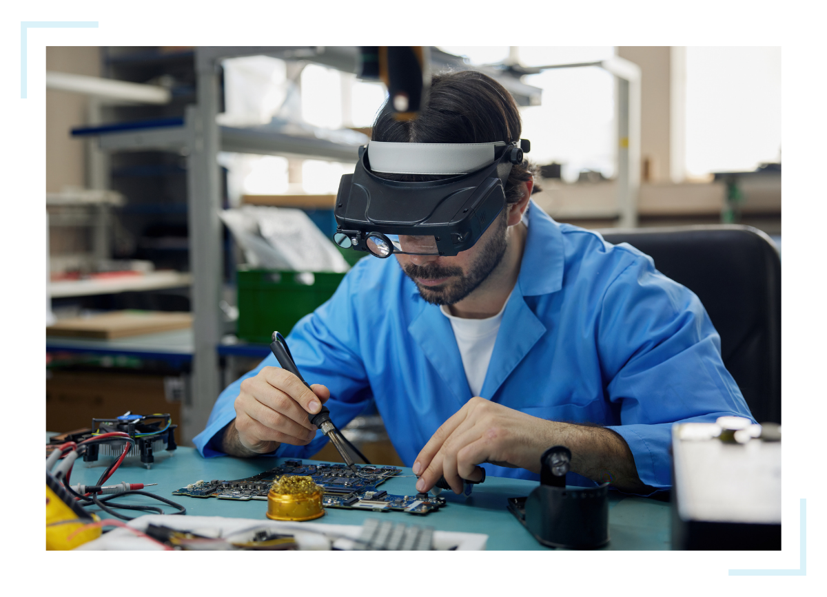A man wearing blue solders an element onto a circuit board in the design stage of turnkey electronic manufacturing.