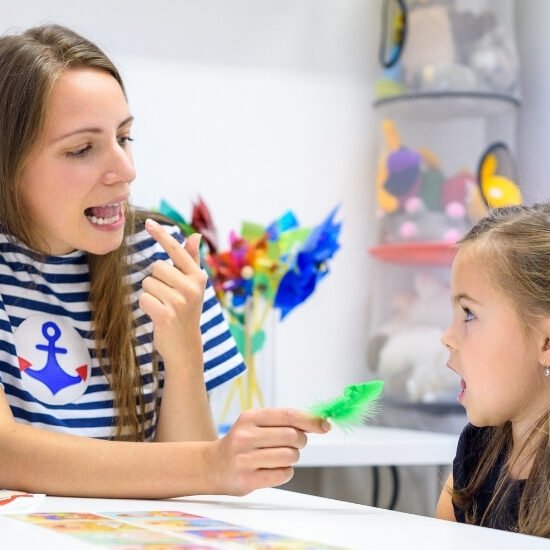 A female speech therapist working with a young girl on tongue placement in a classroom full of colorful items.