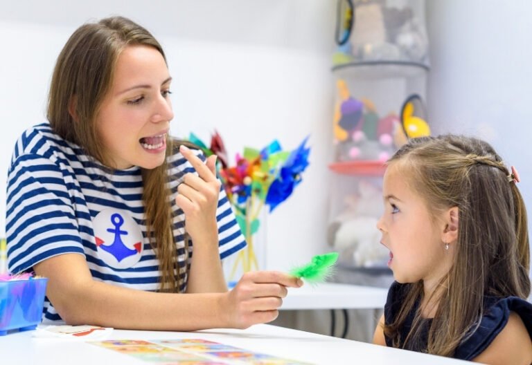 A female speech therapist working with a young girl on tongue placement in a classroom full of colorful items.