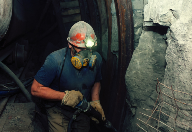 A man working in a mine wearing a hard hat with a light on it and a respirator mask. His work is made safer through tools created by advanced electrical engineering.