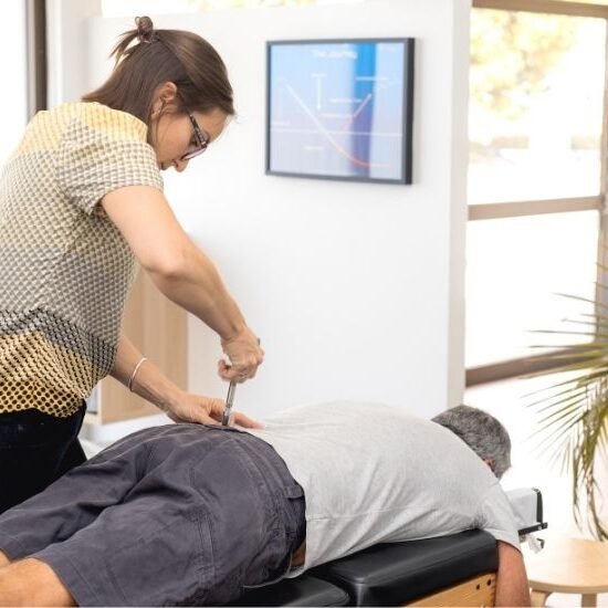 A female chiropractor adjusts a patient who is laying face down on a table using a tool made possible through electronics engineering.
