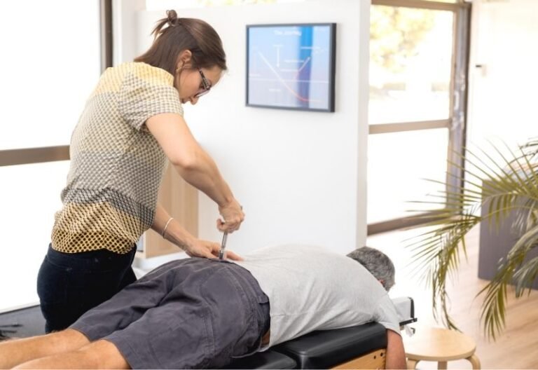 A female chiropractor adjusts a patient who is laying face down on a table using a tool made possible through electronics engineering.