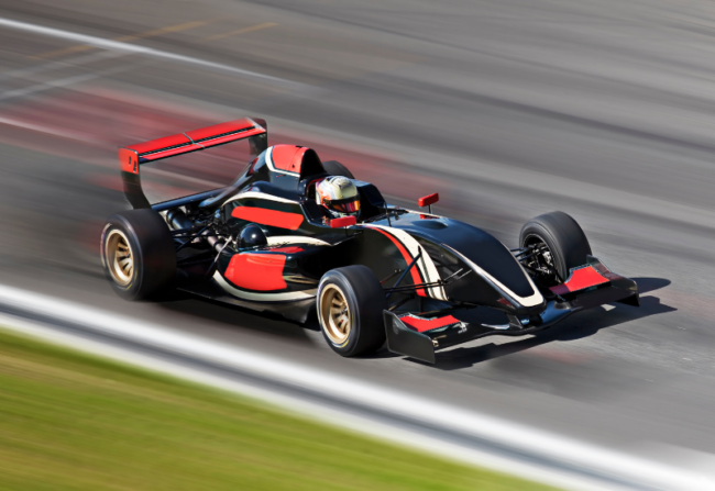 A red and black race car is in focus while the track around it is blurry, showing the care is moving very fast.